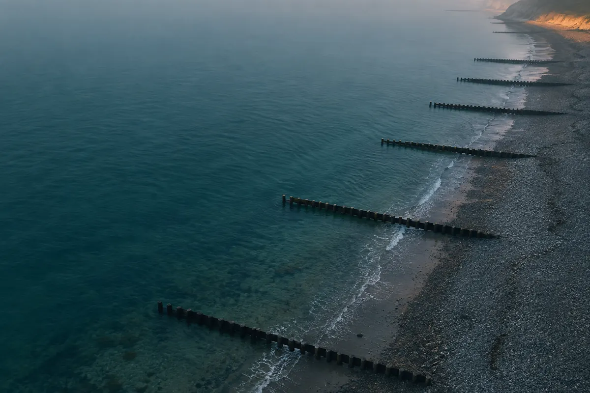 Pebble beach at low tide seen from above at dawn, with wooden breakwaters of alternating lengths and the waterline cutting one halfway.