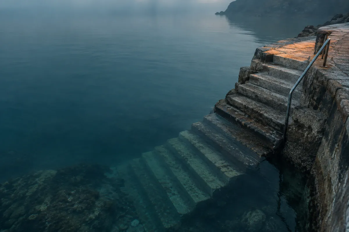 Stone steps descending from a quay into calm water at dawn, dry at the top, half wet in the middle, submerged at the bottom.